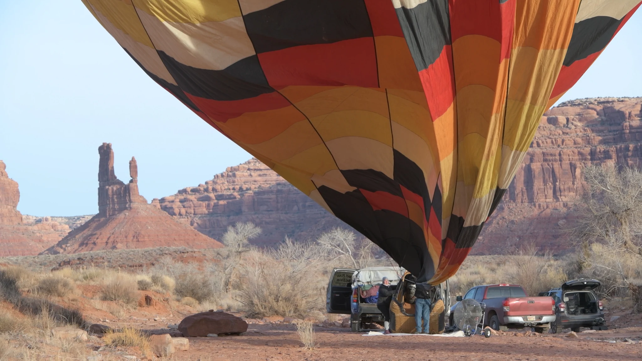 A balloon envelope lifting above a crew and vehicles beneath red cliffs.
