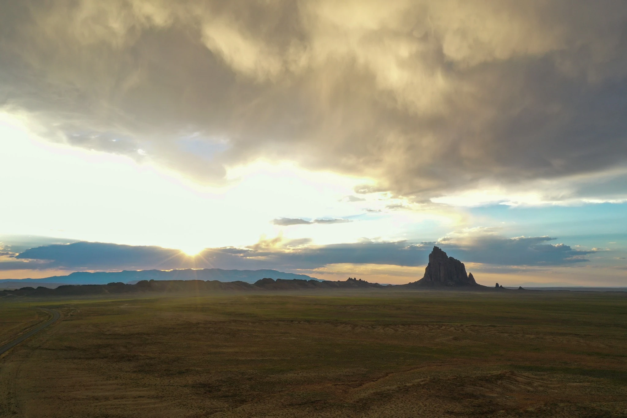 A wide view of Shiprock beneath golden storm clouds, late sunlight striking the desert plain.
