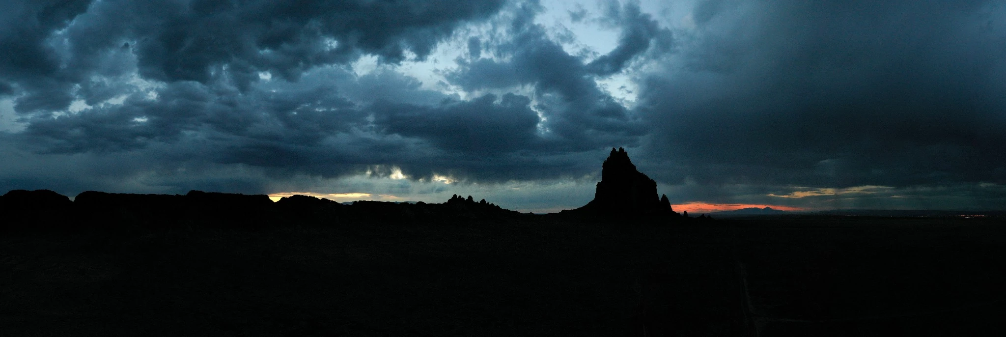A darkened panoramic desert landscape with Shiprock silhouetted below approaching storm clouds.
