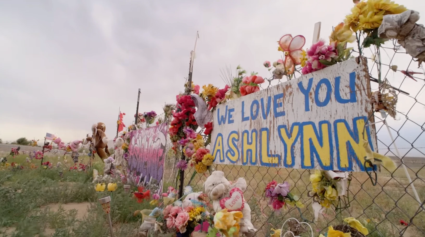 Flowers, toys, and a hand-painted remembrance sign on a roadside fence honoring Ashlynne Mike.