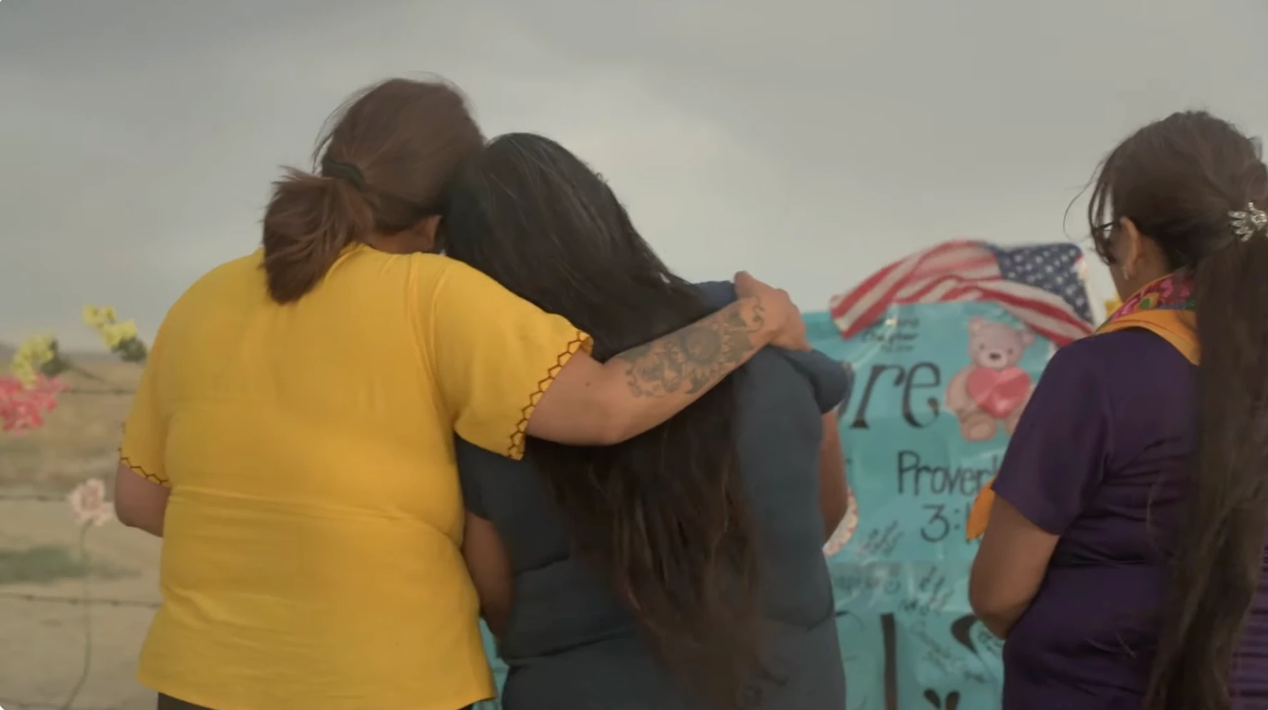 Three women supporting each other at a roadside memorial.