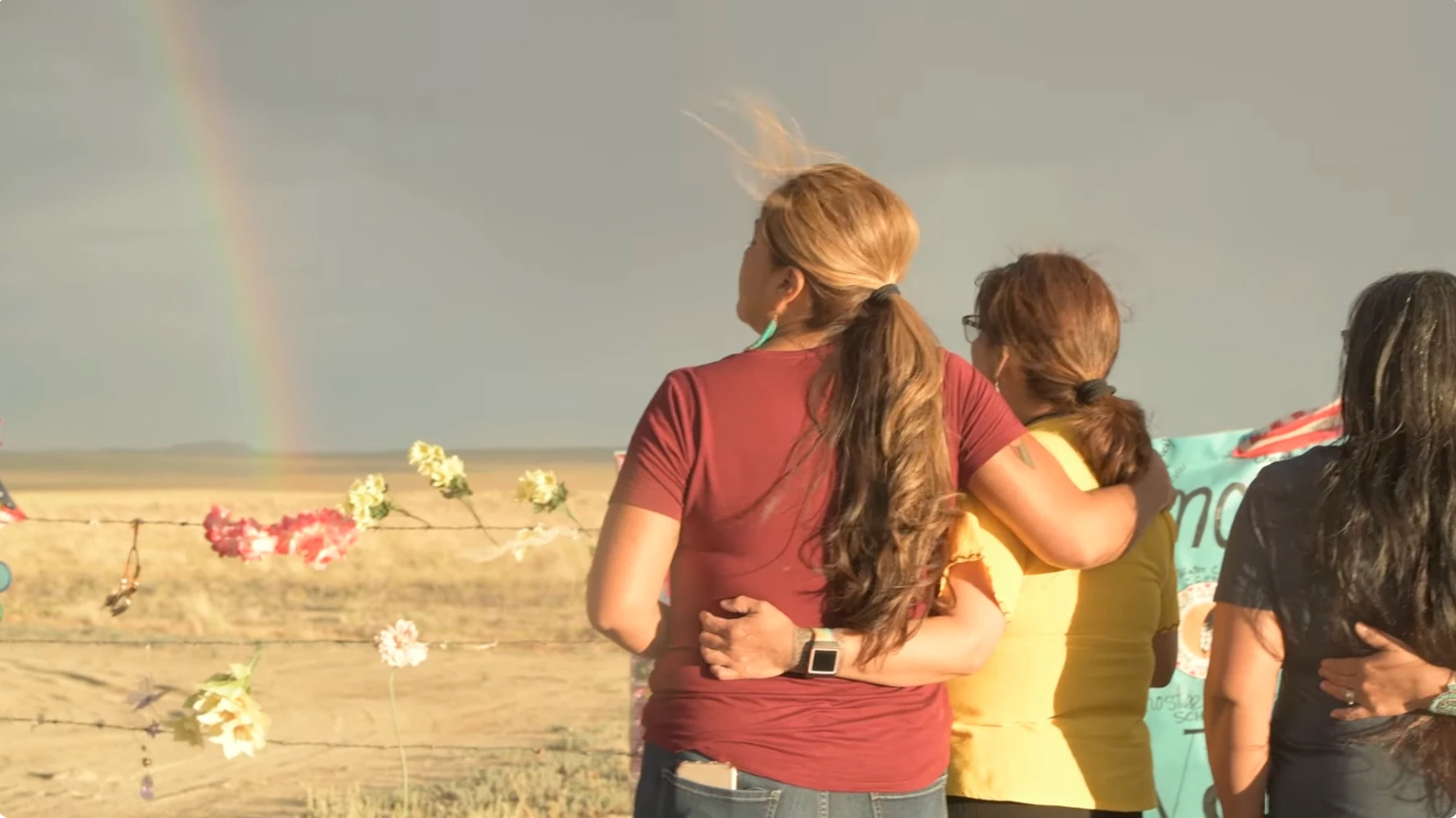 Women standing together, arms around each other, facing the desert as a rainbow emerges on the horizon.